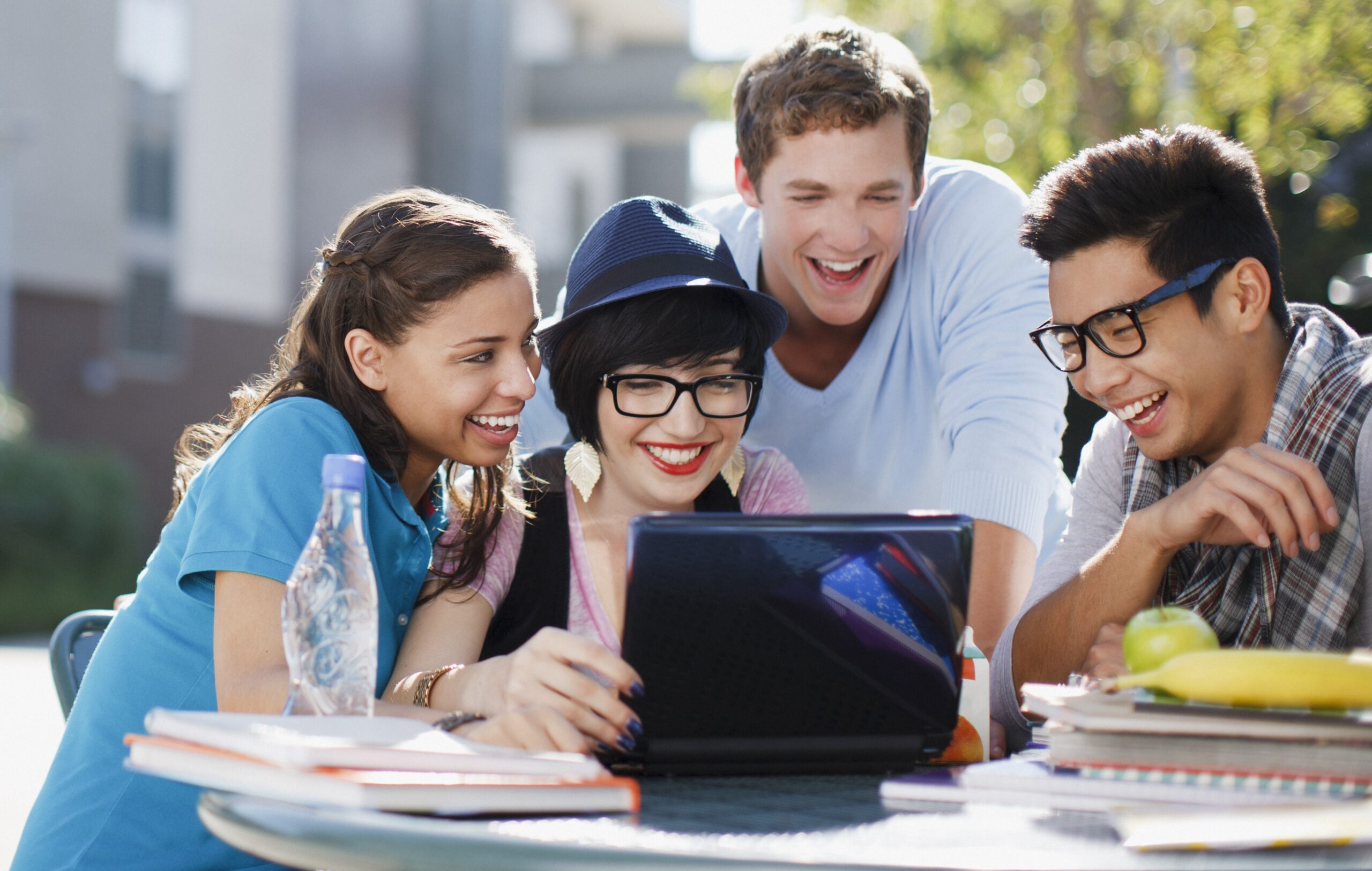 Four students all looking at a laptop screen. Around a table that is outside.